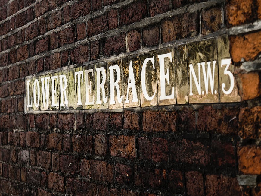A close-up view of a brick wall displaying a street sign with white lettering on a partly green and brown tiled background that reads 'LOWER TERRACE NW3'. The sign is positioned on the left side of th